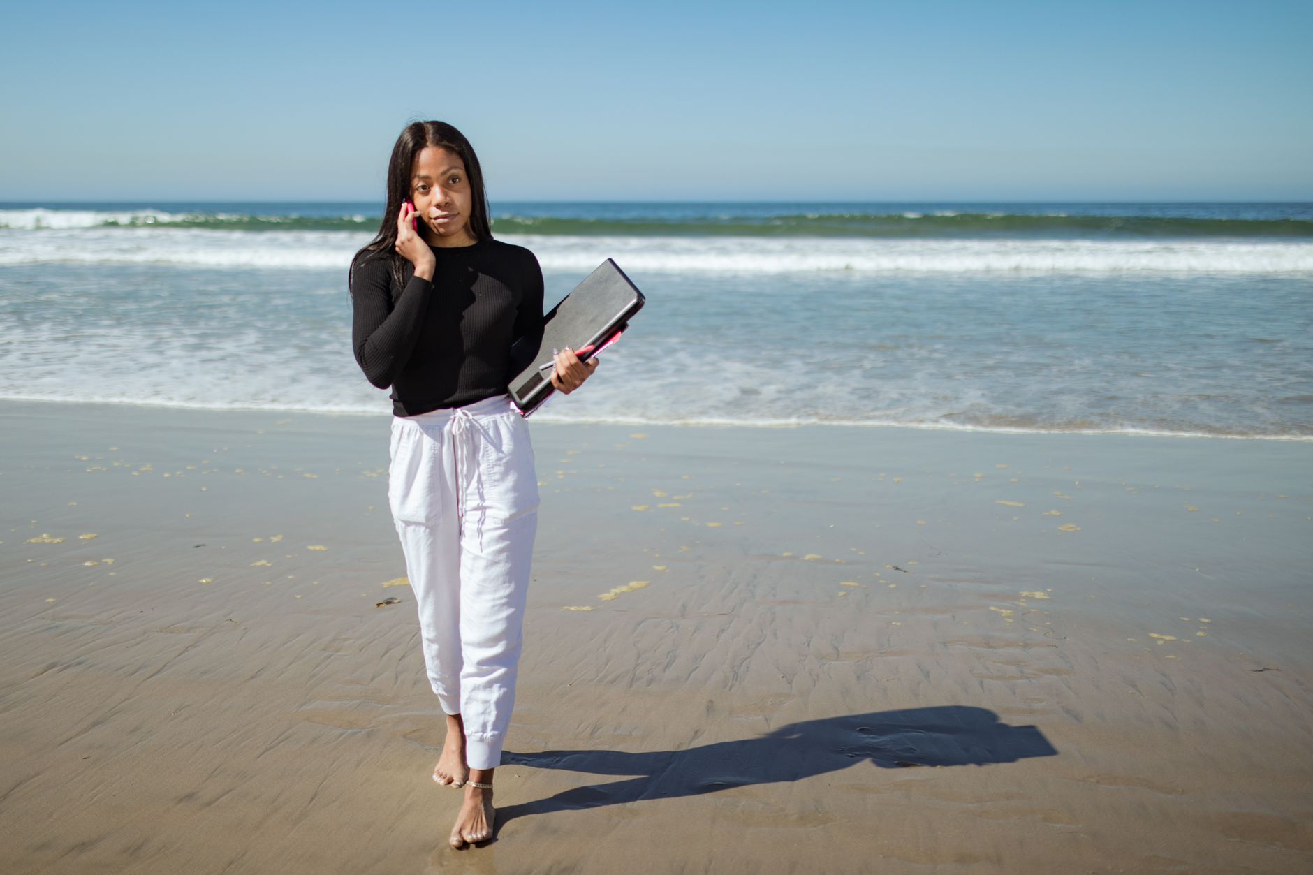 A woman works remotely on the beach, balancing a phone call with her laptop.
